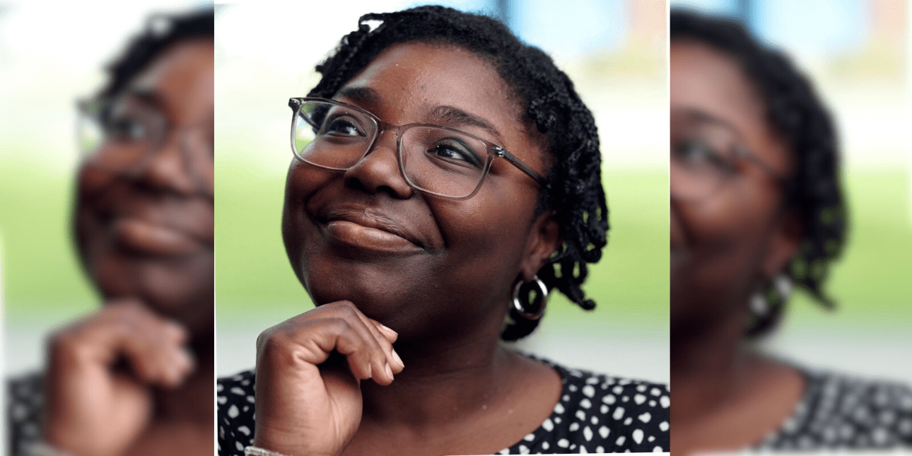 Hannah Abban holding her hand under her chin while looking reflective. Hannah is a Black woman with glasses, short dark hair and wears a white polka dot black top